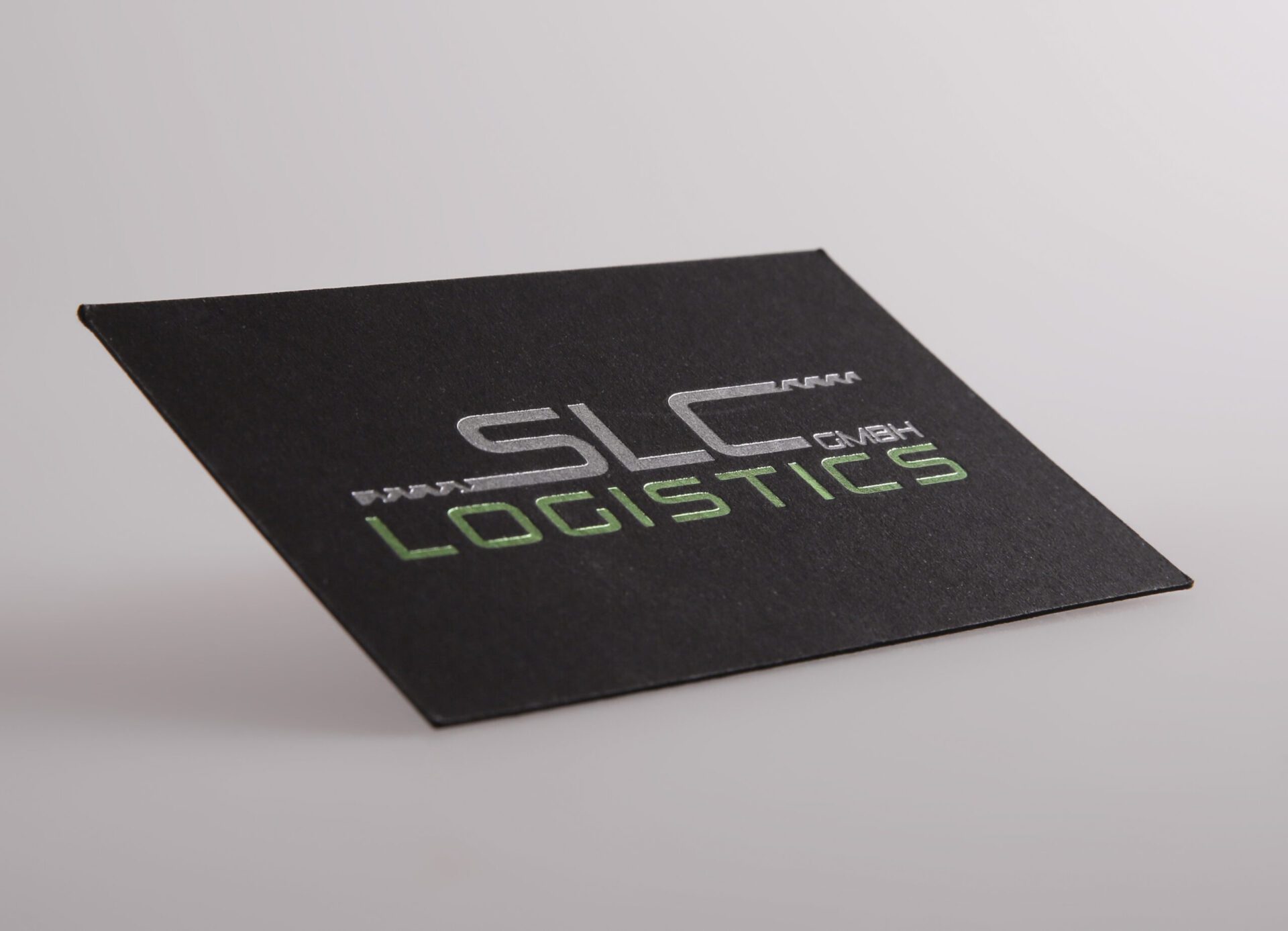 Schwarze Visitenkarte mit glänzender Heißfolienprägung in Silber und Grün mit dem Text „SLC LOGISTICS GmbH“.