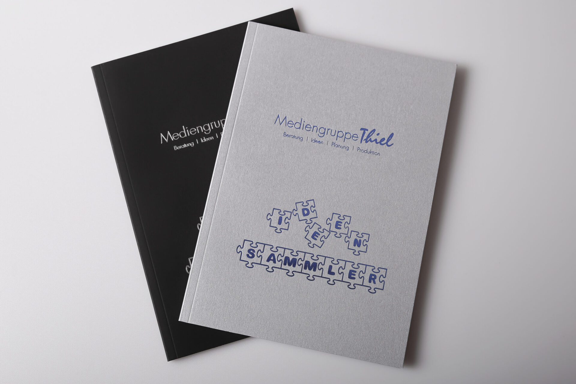 Zwei Notizbücher mit Heißfolienprägung, silbergrau und schwarz, Mediengruppe Thiel, Puzzle-Motiv mit dem Wort Ideensammler.