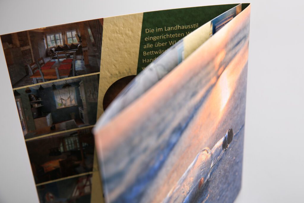 Teilgeöffnete Broschüre mit Titelmotiv einer Flaschenpost am Strand; sichtbar sind Innenaufnahmen einer Ferienwohnung im Landhausstil sowie beschreibender Text zur Ausstattung.