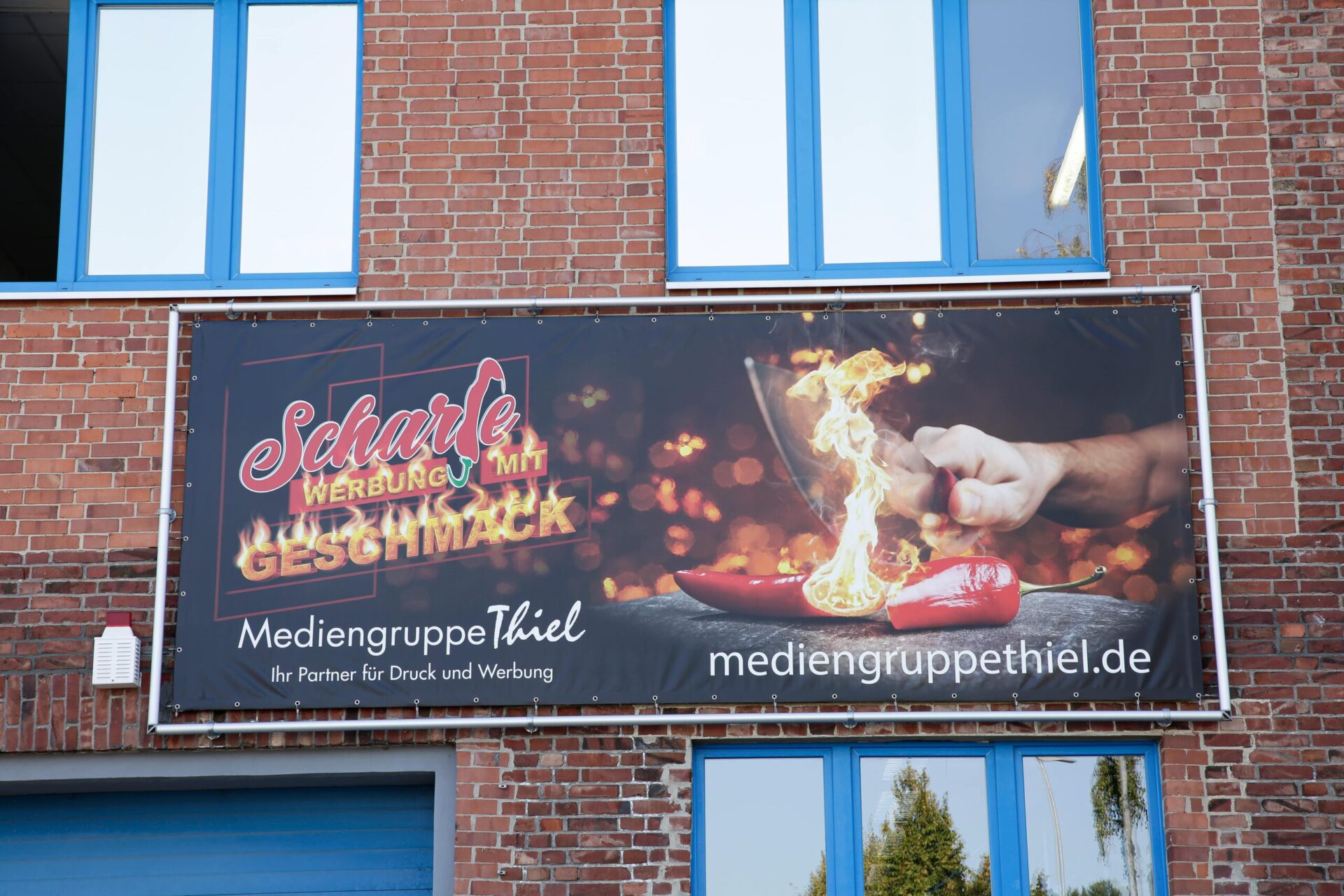 Werbebanner „Scharfe Werbung mit Geschmack“ – Mediengruppe Thiel Außenbanner mit dem Slogan „Scharfe Werbung mit Geschmack“ von der Mediengruppe Thiel, feuriges Design mit einer Hand, die eine scharfe Chilischote schneidet, montiert an einer roten Backsteinfassade.