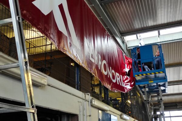 Montage eines großen roten Werbebanners mit der Aufschrift „FERRONORDIC“ durch zwei Monteure auf einer Hubarbeitsbühne in einer Industriehalle.