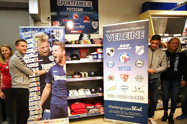 Menschen posieren mit Sponsoring-Banner vor Vereinsmerchandise im Supermarkt