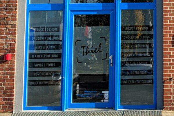 Eingangstür und Fenster mit Glasdekorfolie und Schriftzug „Thiel“ sowie Leistungsangebot, eingerahmt von einer blauen Türrahmenkonstruktion und roter Klinkerfassade.