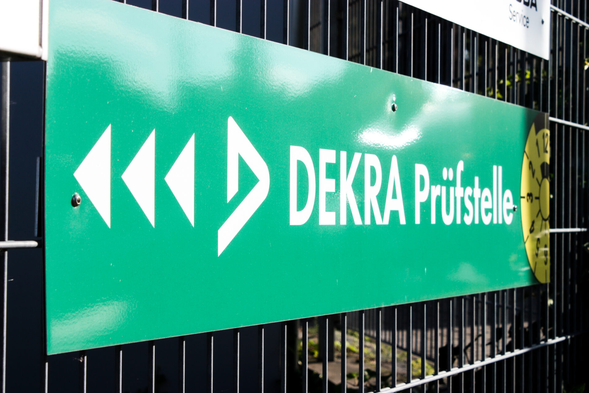 mgt_homepage-117 Grünes Hinweisschild mit DEKRA-Logo und der Aufschrift „DEKRA Prüfstelle“, montiert an einem Metallzaun, mit Richtungspfeilen und TÜV-Plakette zur Orientierung.