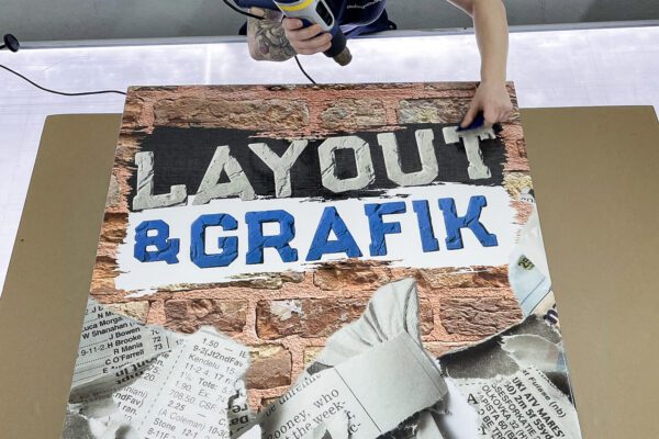 Bedrucktes Schild mit der Aufschrift „LAYOUT & GRAFIK“, das auf einem Arbeitstisch mit Heißluftfön und Rakel bearbeitet wird.