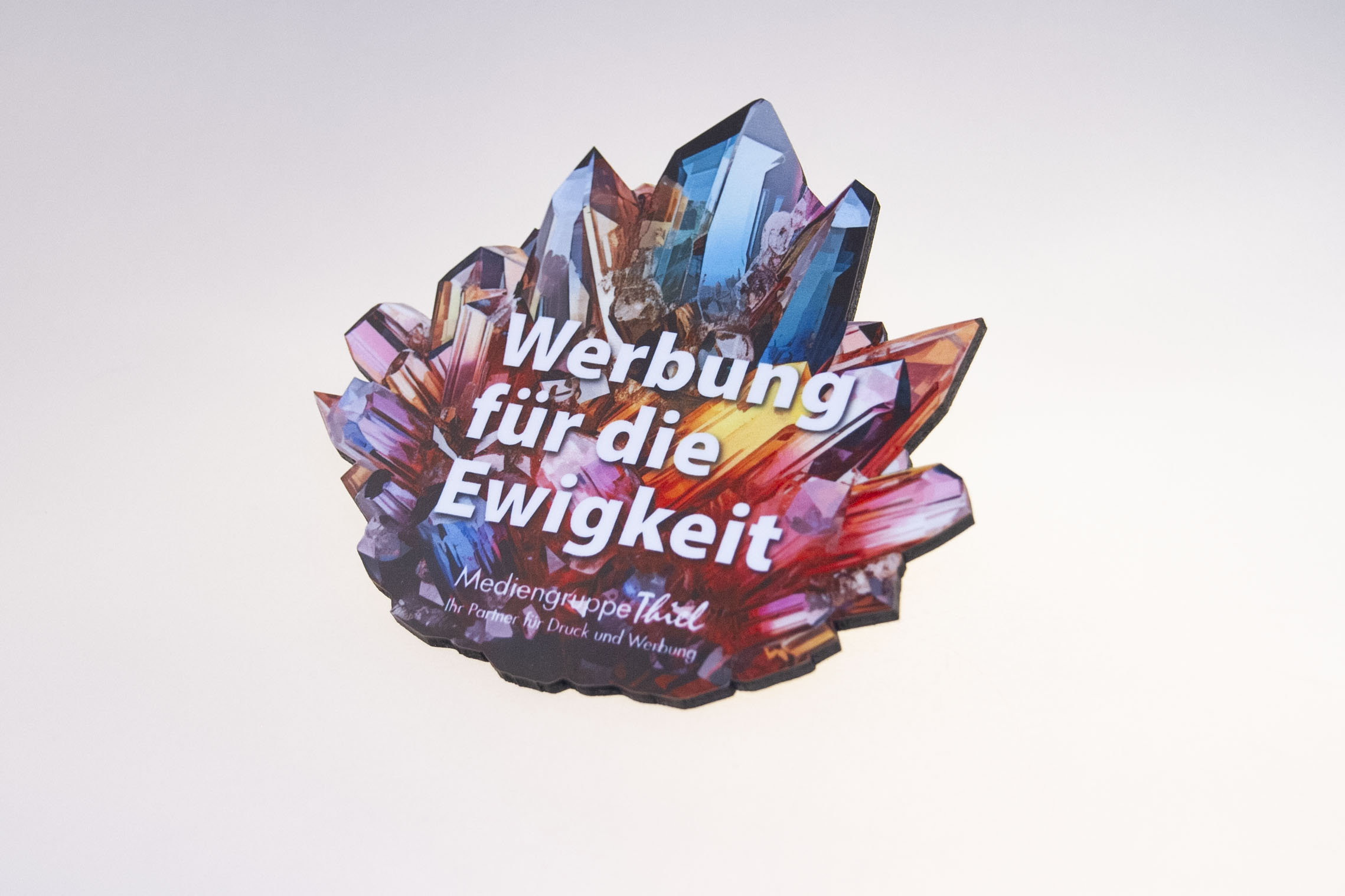Bunter Kristall-Untersetzer mit der Aufschrift „Werbung für die Ewigkeit“ und dem Logo der Mediengruppe Thiel.