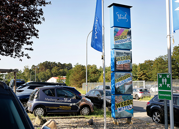 Parkplatz mit Autos und einem Pylonen mit verschiedenen Logos