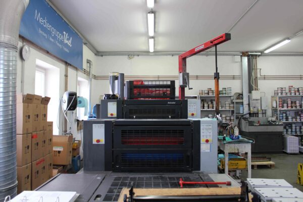 Moderne Offsetdruckmaschine in einer Druckerei, umgeben von Farbdosen, Kartons und Arbeitsmaterialien, mit einem Schild der Mediengruppe Thiel im Hintergrund