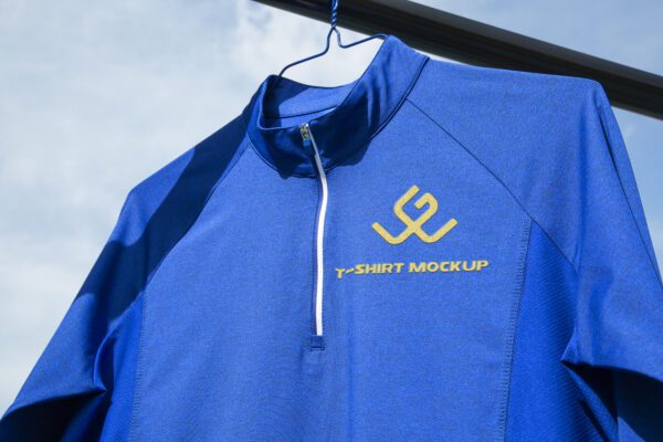 Blaues Funktionsshirt mit gelbem Logo und Schriftzug 'T-Shirt Mockup', hängend an einem Kleiderbügel vor blauem Himmel.