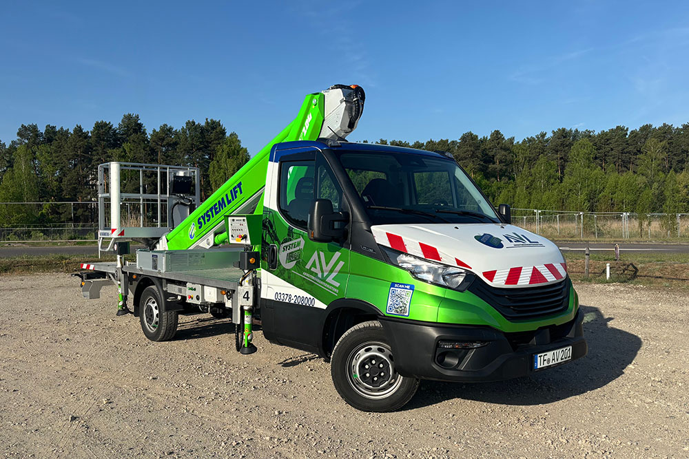 AVL Arbeitsbühne auf Iveco-Fahrgestell mit SystemLift-Aufbau LKW-Arbeitsbühne mit grüner SystemLift-Beschriftung auf Schotterplatz, bereit für den Einsatz, bei sonnigem Wetter vor bewaldetem Hintergrund.