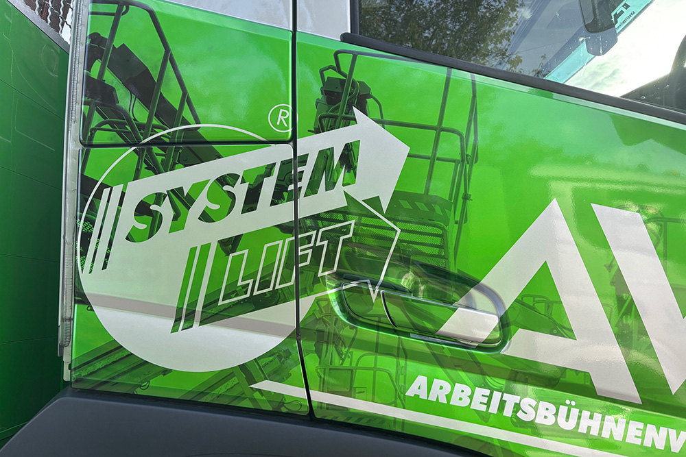 Teilfolierung_LKW_DetailTuer_AVL_web Nahaufnahme der Fahrzeugtür mit SYSTEM LIFT-Logo und AVL-Folierung auf grünem Untergrund.