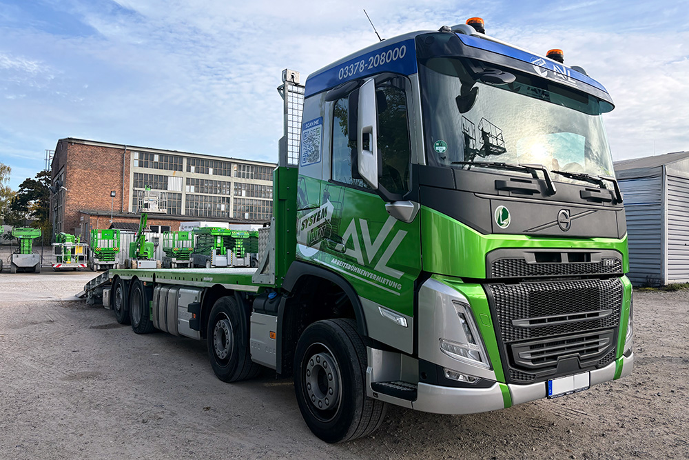 Teilfolierung Sattelschlepper mit Tieflader – AVL Arbeitsbühnenvermietung Großer AVL-Sattelschlepper mit Tieflader und SYSTEM LIFT-Folierung auf einem Firmengelände mit mehreren Hubarbeitsbühnen im Hintergrund.
