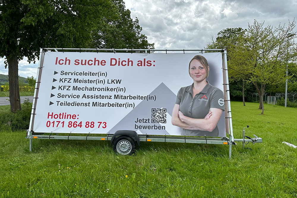 Werbeanhaenger_Stellenanzeige Mobiler Werbeanhänger auf einer Wiese mit großem Banner zur Personalgewinnung. Links steht in großer roter Schrift „Ich suche Dich als:“ gefolgt von fünf aufgelisteten Jobangeboten im KFZ-Bereich. Unten links steht die Hotline-Nummer. Rechts zeigt ein Farbfoto eine Frau in grauem Firmenpolo mit verschränkten Armen. Daneben ein QR-Code mit dem Text „Jetzt bewerben“.