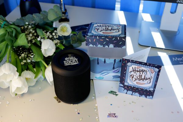 Geburtstags-Arrangement auf Schreibtisch mit Blumenstrauß, Geschenkbox, „Happy Birthday“-Karte, Bluetooth-Lautsprecher und buntem Konfetti.