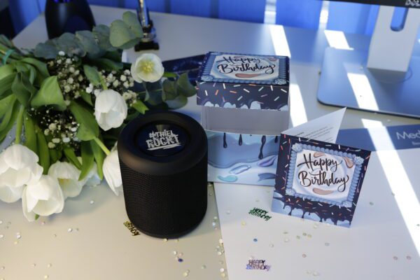 Geburtstags-Geschenkebox mit „Happy Birthday“-Karte, offene Box und schwarzem Mini-Lautsprecher; weiße Tulpen und Konfetti auf dem Schreibtisch.