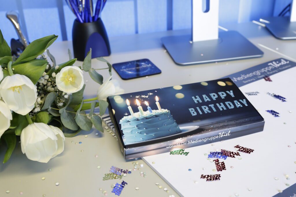 Geschenkebox „HAPPY BIRTHDAY“ mit Torte und Kerzen auf Schreibtisch, weißer Tulpenstrauß, Konfetti, Schreibutensilien, Mediengruppe-Thiel-Schriftzug.