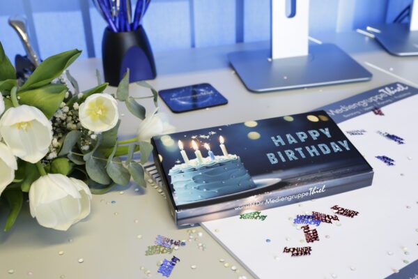 Geschenkebox „HAPPY BIRTHDAY“ mit Torte und Kerzen auf Schreibtisch, weißer Tulpenstrauß, Konfetti, Schreibutensilien, Mediengruppe-Thiel-Schriftzug.