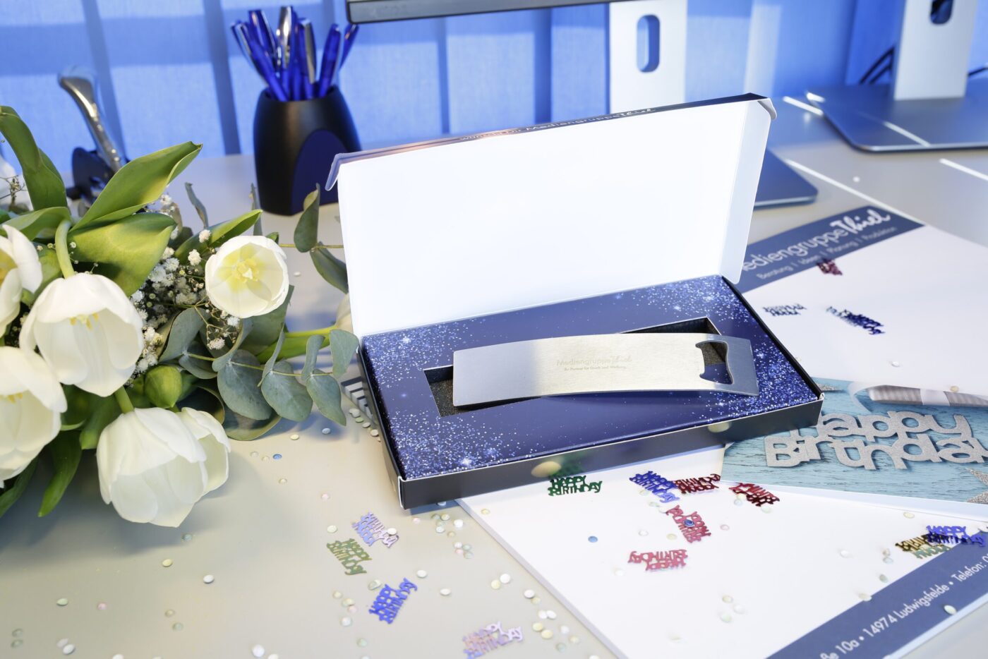 Geschenkebox Mitarbeiterbindung: Offene Box mit Geschenk Geöffnete Geschenkebox mit Einsatz und metallischem Flaschenöffner; links weißer Tulpenstrauß, Konfetti auf Schreibtisch, Büro-Setting.