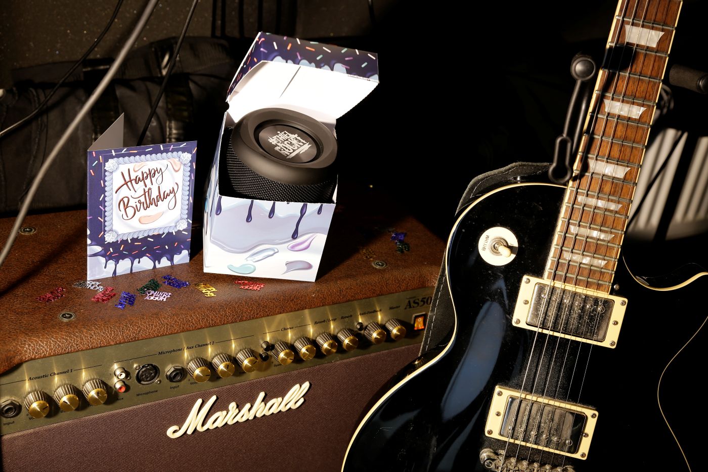 Geschenkebox Mitarbeiterbindung: Musik-Edition Offene Geschenkebox mit kleinem Lautsprecher und Geburtstagskarte auf Marshall-Verstärker; rechts E-Gitarre, Konfetti „Happy Birthday“.