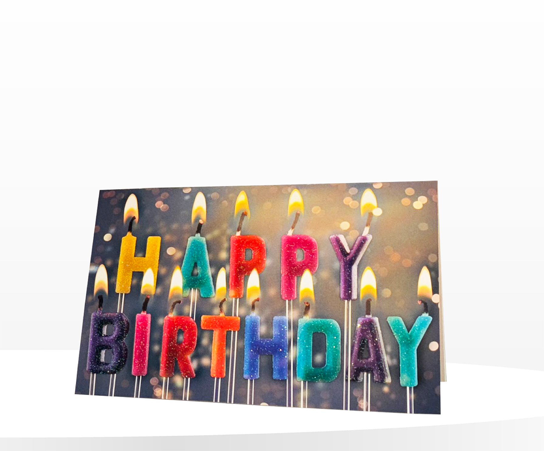 Klappkarte mit brennenden Kerzen in Form der bunten Buchstaben „HAPPY BIRTHDAY“, funkelnder Glitzerlack vor bokehhaftem Hintergrund.