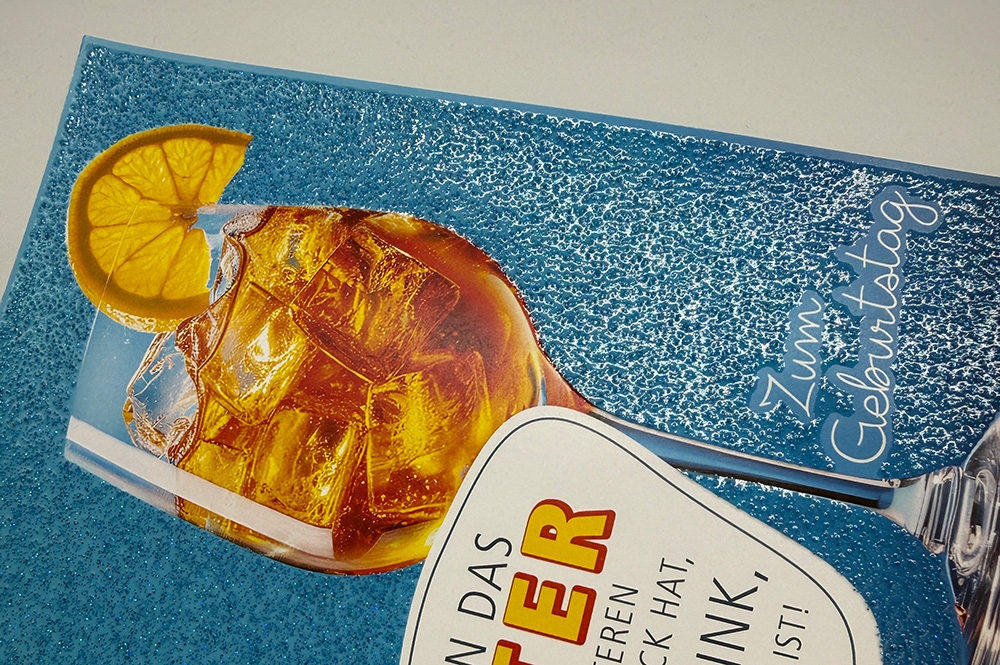 Grußkarte mit Cocktailglas und blauem Glitzerlack-Hintergrund, funkelnde Struktur und Schriftzug „Zum Geburtstag“.