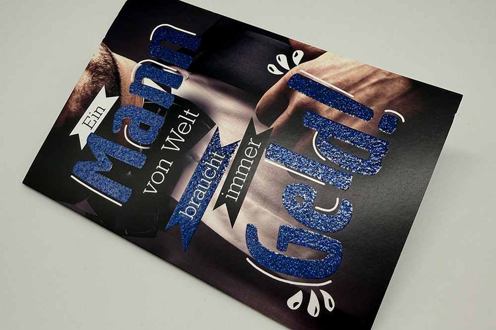 Grußkarte mit blauem Glitzerlack: Text „Ein Mann … braucht immer Geld!“ auf dunklem Motiv, Nahaufnahme mit hochwertigem Effektlack.