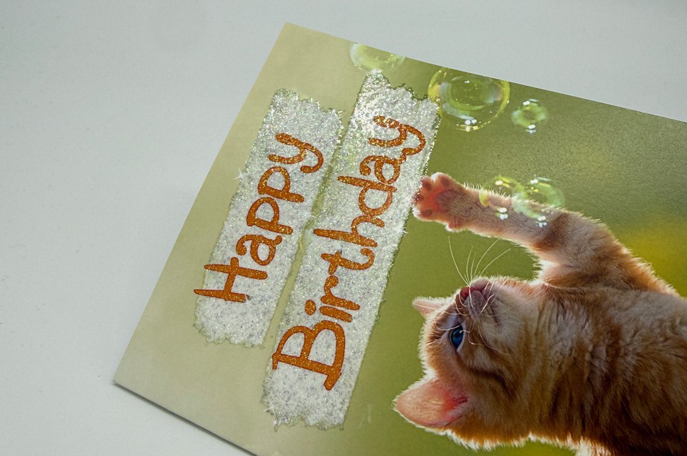 Nahaufnahme einer Geburtstagskarte mit rotem „Happy Birthday“-Schriftzug und weiß funkelndem Glitzerlack; Katze spielt mit Seifenblasen.