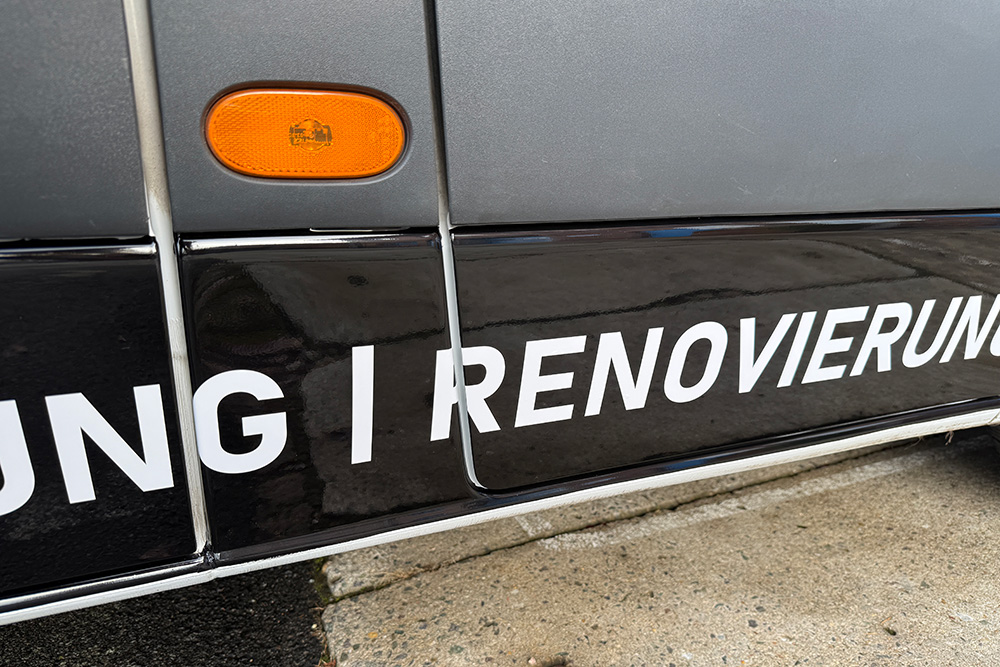 Detail einer Fahrzeugbeschriftung mit weißer Schrift „RENOVIERUNG“ über Türausschnitten und Seitenleuchte auf schwarzer Folie.