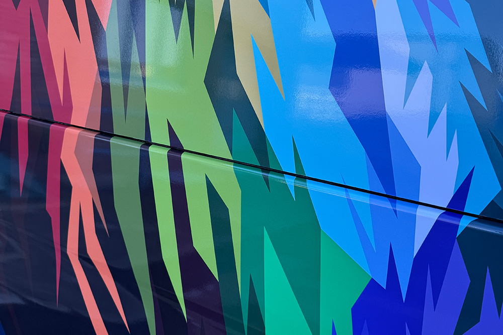 Detail einer bunten Fahrzeugfolie mit abstrakten, zackigen Farbflächen in Blau, Grün und Rot auf einem Transporter.