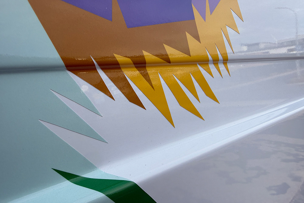 Detailaufnahme einer Fahrzeugfolie mit spitz zulaufenden Farbflächen in Gelb, Grün und Pastell auf weißem Transporterblech.