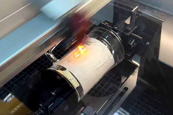 Glasbecher im Lasersystem auf Rotationsachse; der Laser graviert ein Logo umlaufend durch Schutzpapier hindurch.