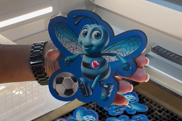 Hand präsentiert blau bedruckten Maskottchen-Untersetzer mit Fußball; frisch aus dem Laser, präzise Kanten, UV-Druck.